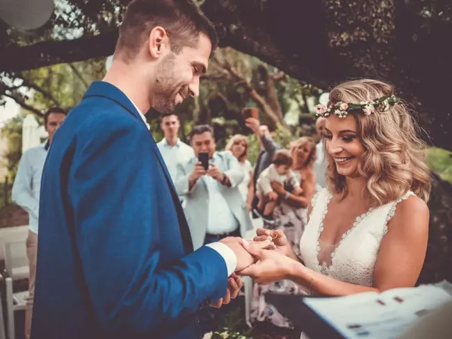 Eine Braut und ein Bräutigam tauschen während ihrer Hochzeit auf Mallorca lächelnd die Ringe aus. Die Gäste im Hintergrund schauen zu und machen Fotos. Die Braut trägt ein Kleid mit Blumenkrone und Spitze, der Bräutigam einen blauen Anzug.