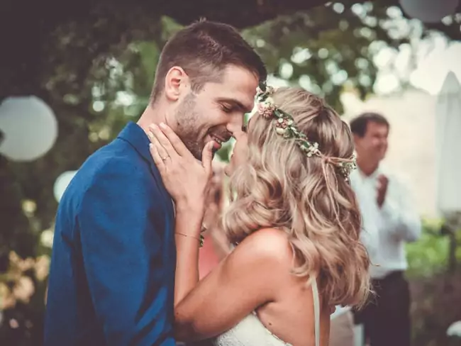 Ein Paar umarmt sich eng und lächelt mit geschlossenen Augen während seiner Hochzeit auf Mallorca. Die Frau trägt eine Blumenkrone und ein weißes Kleid, der Mann einen blauen Blazer. Unscharfe Gäste und üppiges Grün füllen den Hintergrund, eingefangen von einem Hochzeitsfotograf Mallorca.