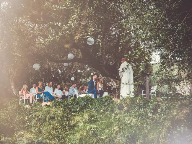 Eine Hochzeit im Freien auf Mallorca entfaltet sich unter großen Bäumen mit hängenden weißen Laternen. Die Gäste sitzen auf weißen Stühlen mit Blick auf das Paar und den Trauzeugen, während das Sonnenlicht durchdringt und eine verträumte Szenerie schafft - perfekt für jeden Hochzeitsfotografen Mallorca zum Festhalten.