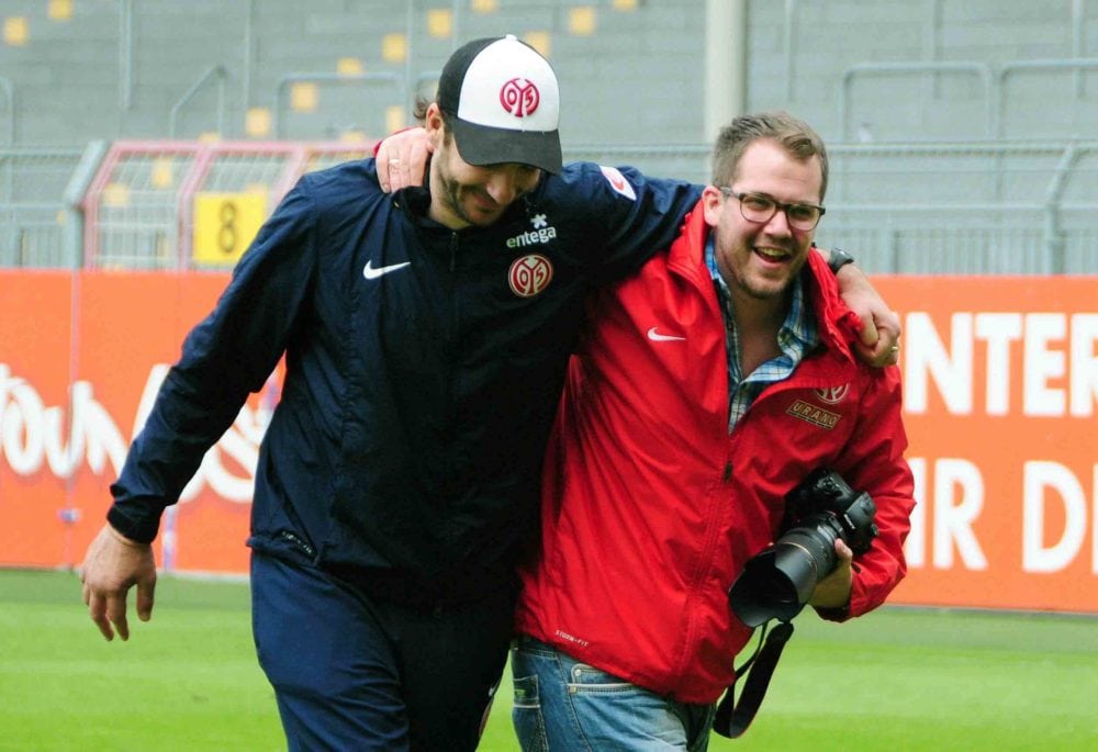 Zwei lächelnde Männer gehen Arm in Arm auf einem Sportplatz; einer trägt eine marineblaue Jacke mit einem Sportlogo, der andere - ein Hochzeitsfotograf Mallorca - trägt eine rote Jacke und eine Brille und hält seine Kamera. Im Hintergrund ist eine Stadionbestuhlung zu sehen.