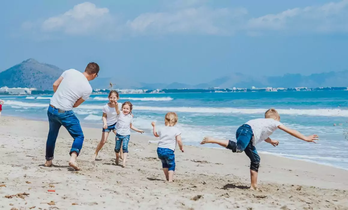 Ein Mann und vier Kinder in legerer Kleidung spielen und rennen zusammen an einem Sandstrand am Meer, mit blauem Himmel und fernen Bergen - perfekte Momente für einen Hochzeitsfotografen Mallorca, um sie während einer Hochzeit auf Mallorca festzuhalten.