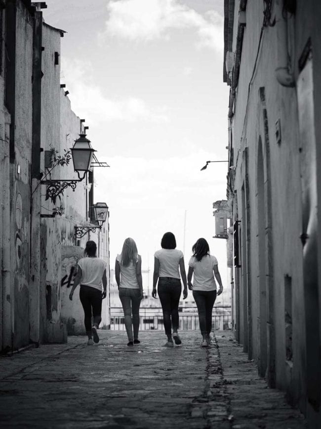Vier Frauen gehen nebeneinander durch eine schmale, mit Steinen gepflasterte Gasse zwischen alten Gebäuden, von hinten gesehen in schwarz-weiß - ein zeitloser Moment, eingefangen von einem Hochzeitsfotografen Mallorca unter einem teilweise bewölkten Himmel.