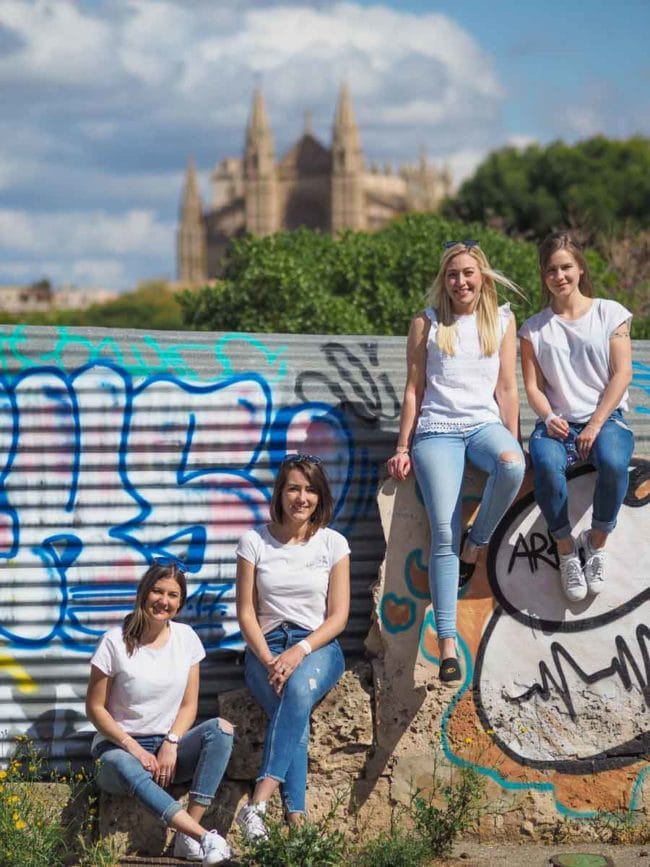 Vier Frauen in weißen Tops und Jeans posieren und lächeln vor einer farbenfrohen Graffiti-Wand, mit einer großen, verschwommenen Kathedrale im Hintergrund - sie fangen einen lustigen Moment während einer Hochzeit auf Mallorca ein, umgeben von üppiger Vegetation.
