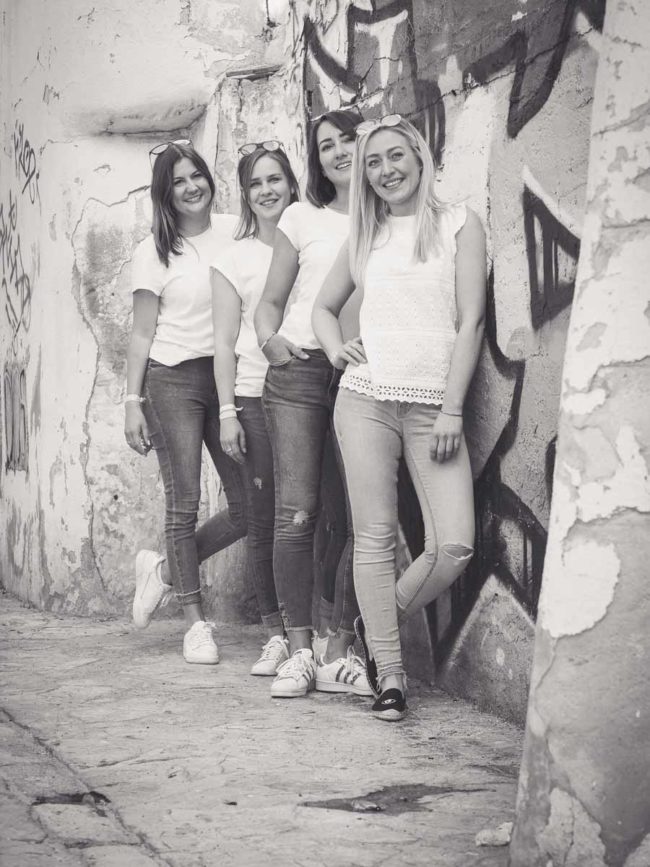 Vier junge Frauen in legerer Kleidung stehen lächelnd in einer Reihe vor einer mit Graffiti beschmierten, verwitterten Wand in einer engen Straße. Aufgenommen von einem Hochzeitsfotograf Mallorca, strahlt das Schwarz-Weiß-Foto Charme und Charakter aus.
