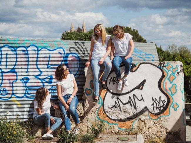 Vier Frauen in weißen Hemden und Jeans sitzen an einer mit Graffiti bedeckten Wand unter einem teilweise bewölkten Himmel und lächeln sich an. Dieser ehrliche Moment fängt die Freude an der Freundschaft ein - die perfekte Inspiration für einen Hochzeitsfotograf Mallorca oder Hochzeit auf Mallorca.