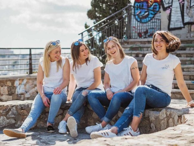 Vier Frauen sitzen auf Steinstufen im Freien, lächeln und unterhalten sich in der Sonne. Ihre legeren Outfits heben sich von einer Graffiti-Wand ab - die perfekte Inspiration für einen Hochzeitsfotografen Mallorca, der auf der Suche nach schönen Momenten vor einer Hochzeit auf Mallorca ist.