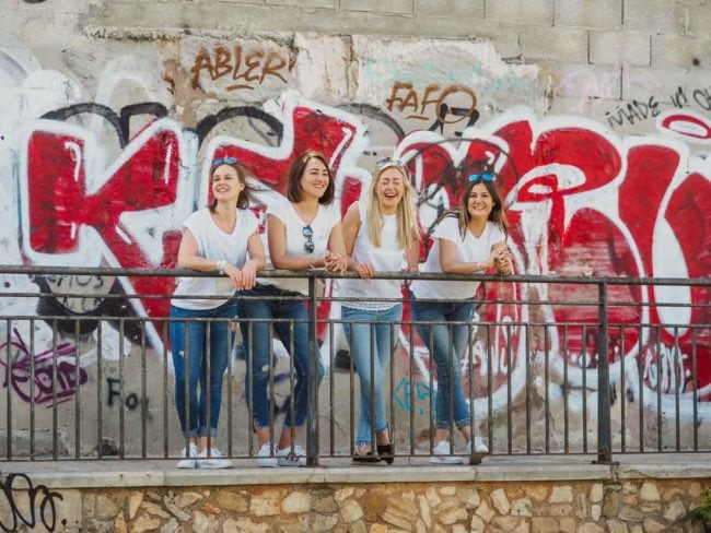 Vier Frauen in weißen Tops und Jeans stehen an einem sonnigen Tag lächelnd an einem Metallgeländer vor einer mit Graffiti bedeckten Wand - die perfekte Inspiration für einen Hochzeitsfotografen Mallorca, der Freundschaften vor einer Hochzeit auf Mallorca festhält.