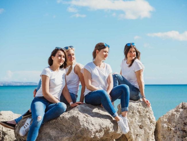 Vier Frauen in weißen Hemden und blauen Jeans sitzen und posieren auf großen Felsen am Meer unter einem strahlend blauen Himmel, lächeln und genießen den sonnigen Tag - und fangen fröhliche Momente ein, die perfekt zu einer Hochzeit auf Mallorca passen.