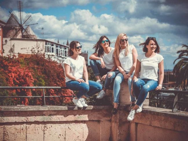 Vier Frauen in legerer Kleidung sitzen auf einer Steinmauer im Freien und lächeln und lachen zusammen während einer Hochzeit auf Mallorca. Im Hintergrund sind eine Windmühle, buntes Laub und ein teilweise bewölkter Himmel zu sehen.