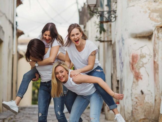 Vier junge Frauen in legeren weißen Oberteilen und Jeans lachen und spielen auf einer schmalen, rustikalen Straße - zwei von ihnen werden Huckepack genommen, während sie alle Freude und Energie ausstrahlen. Perfekte Momente für einen Hochzeitsfotograf Mallorca, um sie vor einer Hochzeit auf Mallorca Feier festzuhalten.