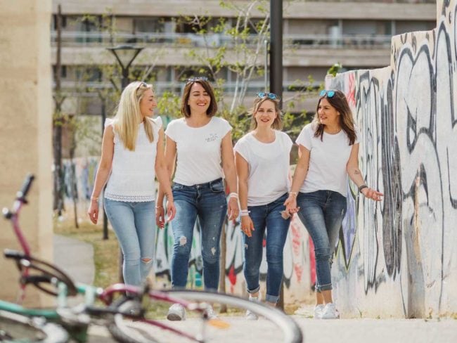 Vier Frauen in weißen Oberteilen und blauen Jeans gehen gemeinsam lächelnd und plaudernd an einer mit Graffiti bedeckten Wand im Freien entlang. Im Vordergrund sind Fahrräder zu sehen - die perfekte Inspiration für einen entspannten Hochzeit auf Mallorca-Moment.