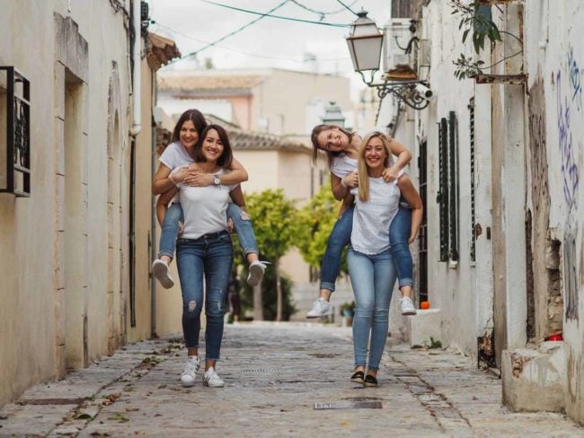 Vier Frauen in legerer Kleidung lachen und reiten Huckepack auf einer sonnenbeschienenen Straße, die von alten Gebäuden und Grünanlagen gesäumt ist - die perfekte Inspiration für einen Hochzeitsfotografen Mallorca, der fröhliche Momente vor einer Hochzeit auf Mallorca festhält.