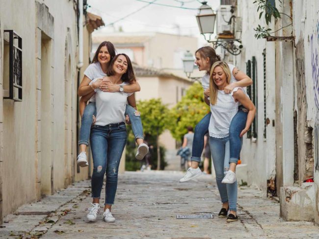 Vier junge Frauen in legerer Kleidung lachen, während zwei von ihnen die anderen auf dem Rücken durch eine sonnenbeschienene Straße tragen, die von weißen Gebäuden und Grün gesäumt ist. Ein talentierter Hochzeitsfotograf Mallorca fängt die fröhlichen Momente ein, die perfekt für eine Hochzeit auf Mallorca sind.
