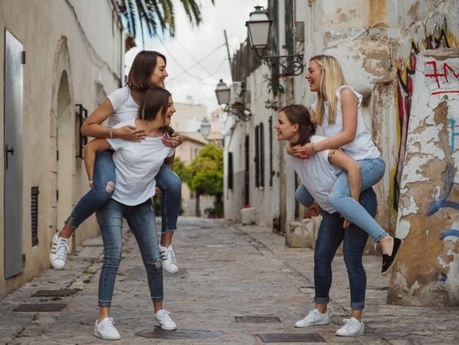 Vier junge Frauen in weißen Oberteilen und Jeans lachen und nehmen sich gegenseitig Huckepack auf einer von alten Gebäuden gesäumten Kopfsteinpflasterstraße - die perfekte Inspiration für einen Hochzeitsfotografen Mallorca oder jeden, der eine Hochzeit auf Mallorca plant.