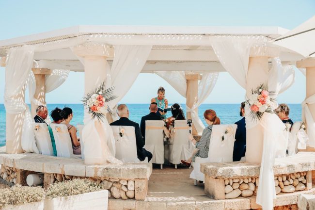 Eine kleine Gruppe nimmt an einer Hochzeit auf Mallorca teil, einer Hochzeitszeremonie im Freien unter einer weißen, mit Blumen geschmückten Pergola, mit dem Meer im Hintergrund. Der Hochzeitsfotograf Mallorca hält die Momente fest, während der Trauzeuge vorne steht und die Gäste nach vorne schauen.