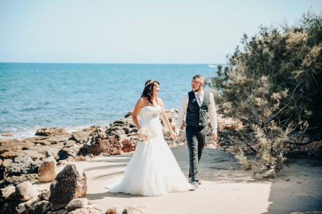 Eine Braut im weißen Kleid und ein Bräutigam mit Weste und Krawatte gehen Hand in Hand einen felsigen Strandweg entlang und lächeln sich an, während im Hintergrund das Meer und der blaue Himmel zu sehen sind - der Zauber einer Hochzeit auf Mallorca.