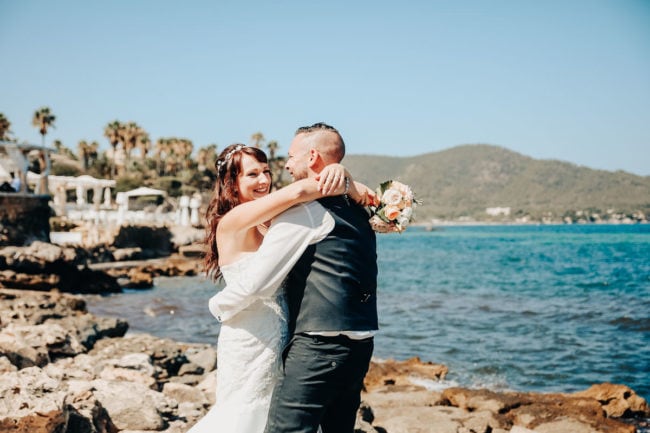 Eine Braut und ein Bräutigam umarmen sich und lächeln freudig an einem felsigen Ufer am Meer, mit klarem blauen Himmel, Palmen und grünen Hügeln im Hintergrund - ein perfekter Moment für Hochzeitsfotograf Mallorca während einer unvergesslichen Hochzeit auf Mallorca.