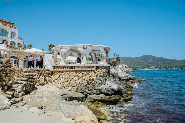 Auf einer Steinterrasse am Meer mit weißem Vorhang findet eine Hochzeit auf Mallorca statt. Die Gäste versammeln sich unter einem Baldachin mit Blick auf die felsige Küste und das blaue Wasser, mit Hügeln und Gebäuden im Hintergrund unter einem klaren blauen Himmel.