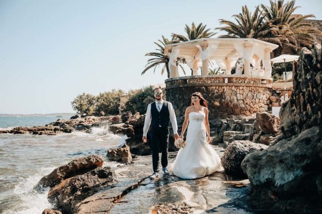 Braut und Bräutigam spazieren Hand in Hand am felsigen Ufer des Meeres entlang, in der Nähe eines weißen Steinpavillons, umgeben von Palmen an einem sonnigen Tag - perfekte Momente für einen Hochzeitsfotografen Mallorca, um Ihre unvergessliche Hochzeit auf Mallorca festzuhalten.