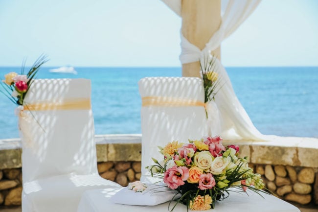 Zwei weiße Stühle mit pfirsichfarbenen Bändern und Blumenschmuck stehen vor einem kleinen Tisch mit einem Strauß rosa und weißer Blumen, draußen am Meer - perfekt für eine Hochzeit auf Mallorca, wunderschön eingefangen von einem Hochzeitsfotografen Mallorca.