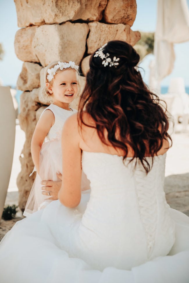 Eine Braut in einem weißen Kleid kniet und lächelt ein junges Blumenmädchen in Rosa mit einem geblümten Stirnband an. Aufgenommen von einem Hochzeitsfotograf Mallorca, teilen sie einen süßen Moment im Freien in der Nähe von Steinsäulen auf ihrer Hochzeit auf Mallorca.