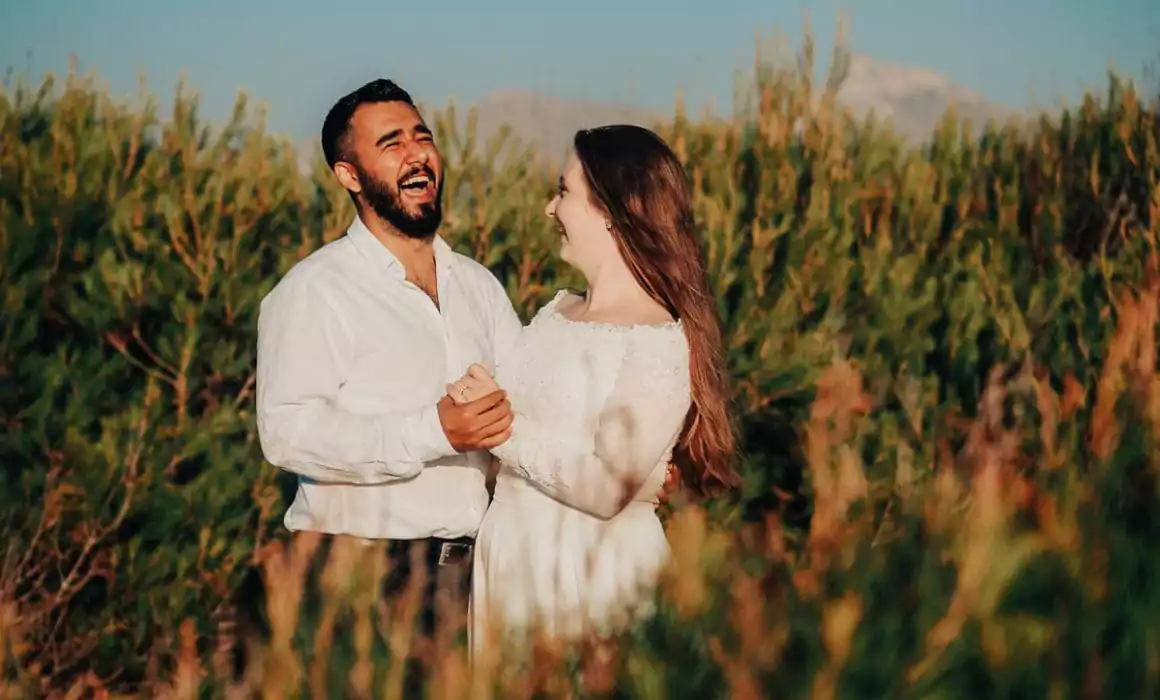 Ein Mann und eine Frau in weißer Kleidung stehen im Freien zwischen hohen grünen Sträuchern, halten sich an den Händen und lachen zusammen unter einem klaren blauen Himmel - die Freude über eine Hochzeit auf Mallorca.