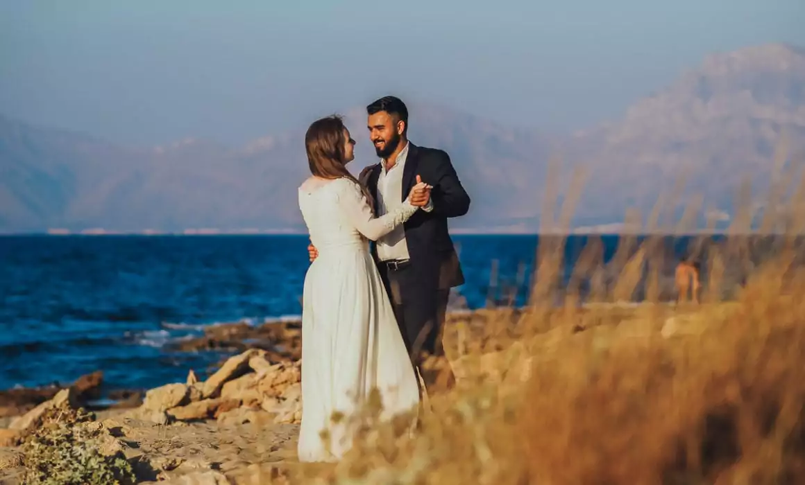 Ein formell gekleidetes Paar tanzt zusammen an einem Sandstrand, mit dem Meer und den fernen Bergen im Hintergrund unter einem klaren blauen Himmel - die perfekte Inspiration für eine Hochzeit auf Mallorca, aufgenommen von einem Hochzeitsfotograf Mallorca.