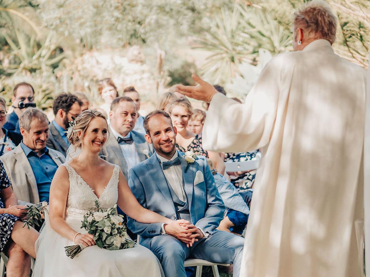 Eine Braut und ein Bräutigam sitzen händchenhaltend und lächeln sich an während ihrer Hochzeit auf Mallorca. Der Zeremonienmeister steht vorne und spricht zu ihnen, während die Gäste und der Hochzeitsfotograf Mallorca von ihren Plätzen im Hintergrund zusehen.