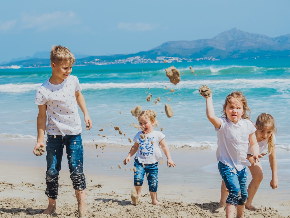 Vier kleine Kinder spielen vergnügt an einem Sandstrand in der Nähe des Meeres, werfen Sand mit Bergen und blauem Himmel im Hintergrund - perfekte Momente für einen Hochzeitsfotografen Mallorca.