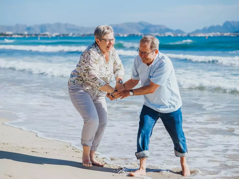 Ein älteres Paar lacht und hält sich an den Händen, während es an einem Sandstrand in der Nähe des Ozeans spielt, mit sanften Wellen und fernen Bergen - wunderschön eingefangen wie von einem Hochzeitsfotografen Mallorca während einer romantischen Hochzeit auf Mallorca.