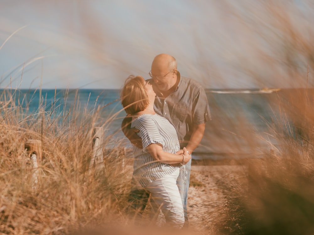 Ein älteres Paar steht eng beieinander, lächelnd und sich umarmend an einem Strand mit hohem Gras, das Meer im Hintergrund unter einem klaren Himmel - eingefangen von einem Hochzeitsfotografen Mallorca, perfekt für Erinnerungen an eine Hochzeit auf Mallorca.