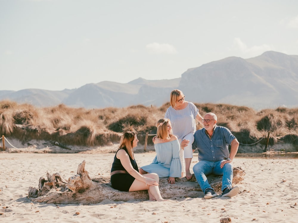 Vier Personen, zwei Männer und zwei Frauen, sitzen und stehen auf einem großen Baumstamm an einem Sandstrand mit Bergen im Hintergrund. Lächelnd in der Sonne, sehen sie so fröhlich aus wie auf einer Hochzeit auf Mallorca, perfekt für jeden Hochzeitsfotograf Mallorca zu erfassen.