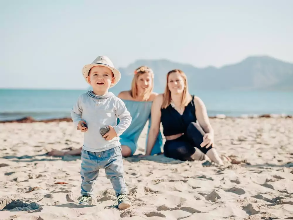 Ein Kleinkind mit Sonnenhut steht lächelnd an einem Sandstrand und hält kleine Steine in der Hand, während zwei Frauen in der Nähe des Ufers sitzen - eine idyllische Szene, perfekt für jeden Hochzeitsfotografen Mallorca oder diejenigen, die eine Hochzeit auf Mallorca planen.