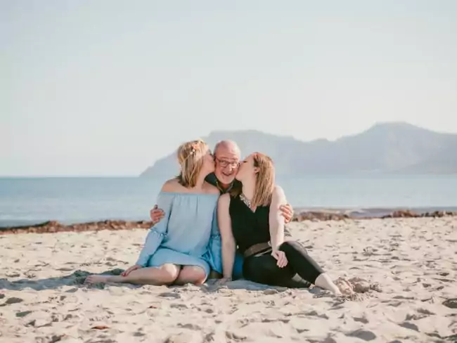 Drei Personen sitzen an einem Sandstrand; zwei Frauen küssen einen älteren Mann auf die Wange. Das Meer und die Berge in der Ferne bilden die Kulisse für einen klaren, sonnigen Tag - perfekt für Erinnerungen, die von einem Hochzeitsfotografen Mallorca eingefangen wurden.