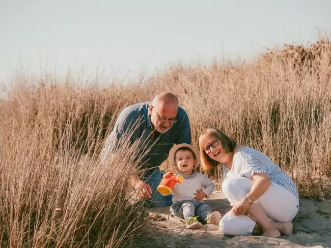 Ein älterer Mann und eine Frau lächeln, während sie im hohen, trockenen Gras neben einem glücklichen Kleinkind knien, das ein Spielzeug hält. Die Gruppe posiert zusammen auf einem sandigen, sonnenbeschienenen Weg im Freien - ein idyllischer Moment, den jeder Hochzeitsfotograf Mallorca perfekt festhalten kann.