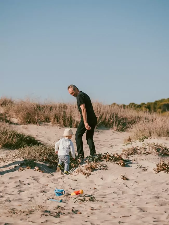 Ein Erwachsener und ein Kind spazieren an einem Sandstrand unter einem klaren blauen Himmel entlang, Spielzeug in der Nähe verstreut. Dieser heitere Moment fängt die Essenz einer Hochzeit auf Mallorca ein, mit trockenem Gras im Hintergrund, das die malerische Szene vervollständigt.
