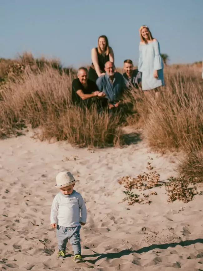 Ein Kleinkind mit Hut steht auf sandigem Boden vor hohem Gras, während fünf Erwachsene im Hintergrund gemeinsam posieren - ein fröhlicher Moment während einer Hochzeit auf Mallorca an einem sonnigen Strandtag.