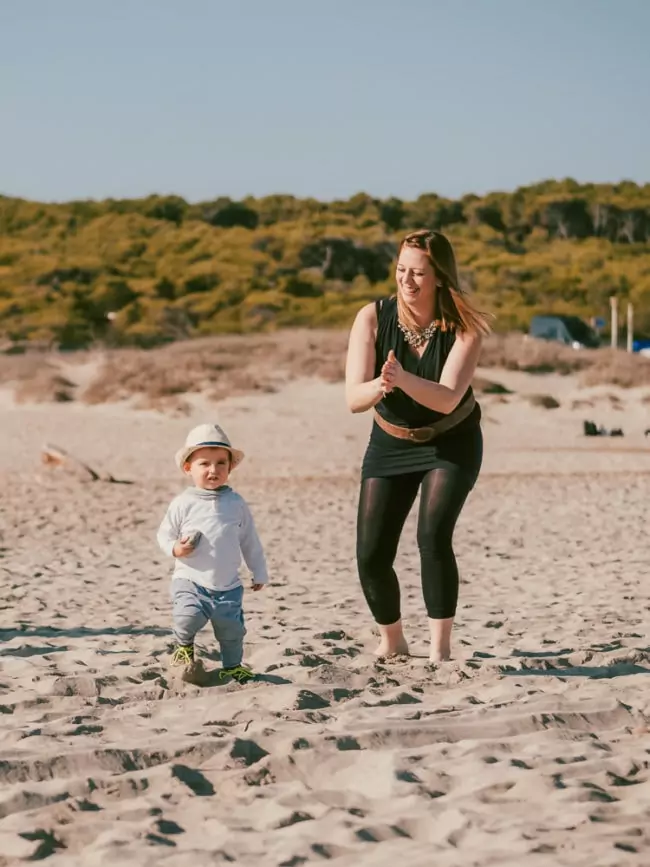 Eine Frau und ein kleines Kind spazieren zusammen an einem Sandstrand unter einem klaren Himmel, mit Grün hinter ihnen. Das Kind lächelt in einen Hut, während die Frau sie ermutigt - eine idyllische Szene, die oft von einem Hochzeitsfotografen Mallorca eingefangen wird.