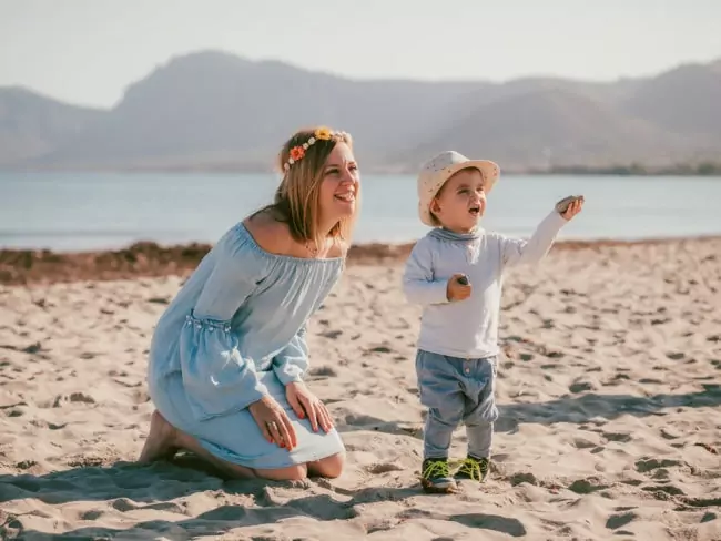 Eine lächelnde Frau in einem hellblauen Kleid und einer Blumenkrone kniet an einem Sandstrand neben einem kleinen Kind, mit den Bergen und dem Wasser im Hintergrund - und fängt den Zauber einer Hochzeit auf Mallorca ein.