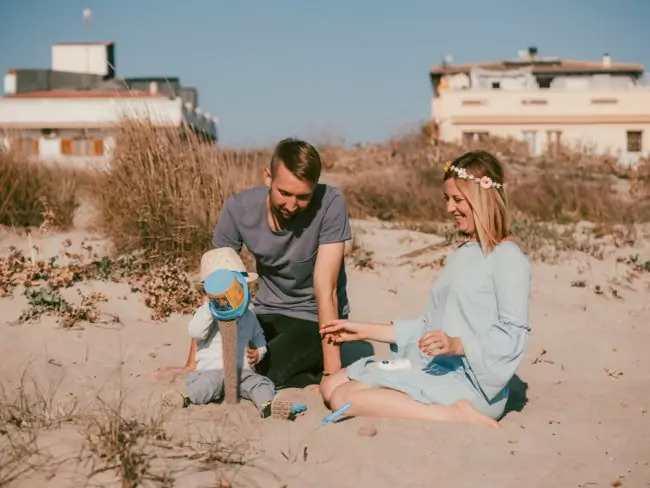 Ein Mann, eine Frau und ein kleines Kind spielen zusammen an einem Sandstrand in der Nähe von getrocknetem Gras, mit Häusern und klarem, blauem Himmel im Hintergrund - ein schöner Moment, perfekt für einen Hochzeitsfotografen Mallorca, der eine Hochzeit auf Mallorca festhält.