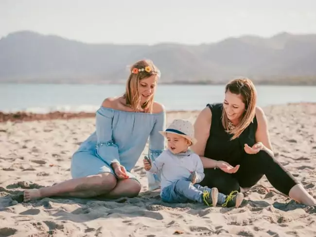Zwei Frauen sitzen an einem Sandstrand mit einem Kleinkind zwischen ihnen, Berge und Wasser im Hintergrund. Dieser zärtliche Moment, perfekt für einen Hochzeitsfotograf Mallorca, fängt die Freude über eine Hochzeit auf Mallorca am Meer ein.