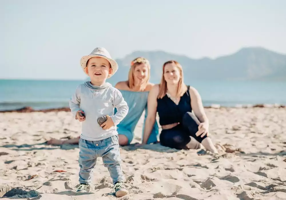 Ein Kleinkind mit Hut steht lächelnd an einem Sandstrand und hält zwei Felsen. Im Hintergrund sitzen zwei Frauen auf dem Sand mit den Bergen und dem Meer im Hintergrund - eine perfekte Szene für jeden Hochzeitsfotograf Mallorca an einem sonnigen Tag.