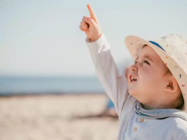 Ein kleines Kind, das einen Sonnenhut und ein helles Hemd trägt, lächelt und zeigt in den Himmel, während es an einem sonnigen Strand auf Mallorca steht - perfekt für das Einfangen fröhlicher Momente durch einen Hochzeitsfotografen Mallorca, mit dem Meer und dem klaren blauen Himmel im Hintergrund.