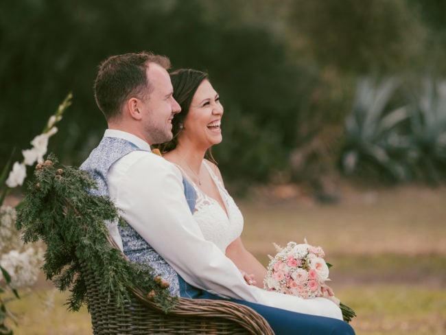 Eine Braut und ein Bräutigam sitzen Seite an Seite auf einer Korbbank im Freien, lächeln und schauen nach links. Die Braut hält einen rosafarbenen und weißen Blumenstrauß in der Hand, während die Bank mit Grünpflanzen geschmückt ist - perfekte Erinnerungen, festgehalten von einem Hochzeitsfotografen Mallorca.