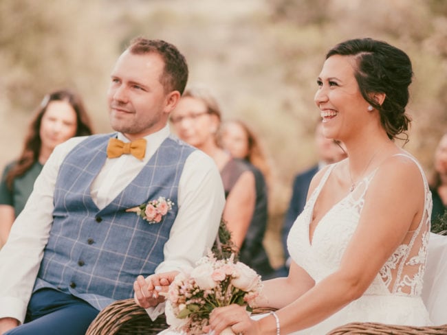 Eine lächelnde Braut in Weiß und ein Bräutigam mit gelber Fliege sitzen Hand in Hand im Freien, umgeben von Gästen während ihrer Hochzeit auf Mallorca - wunderschön eingefangen von einem Hochzeitsfotograf Mallorca.