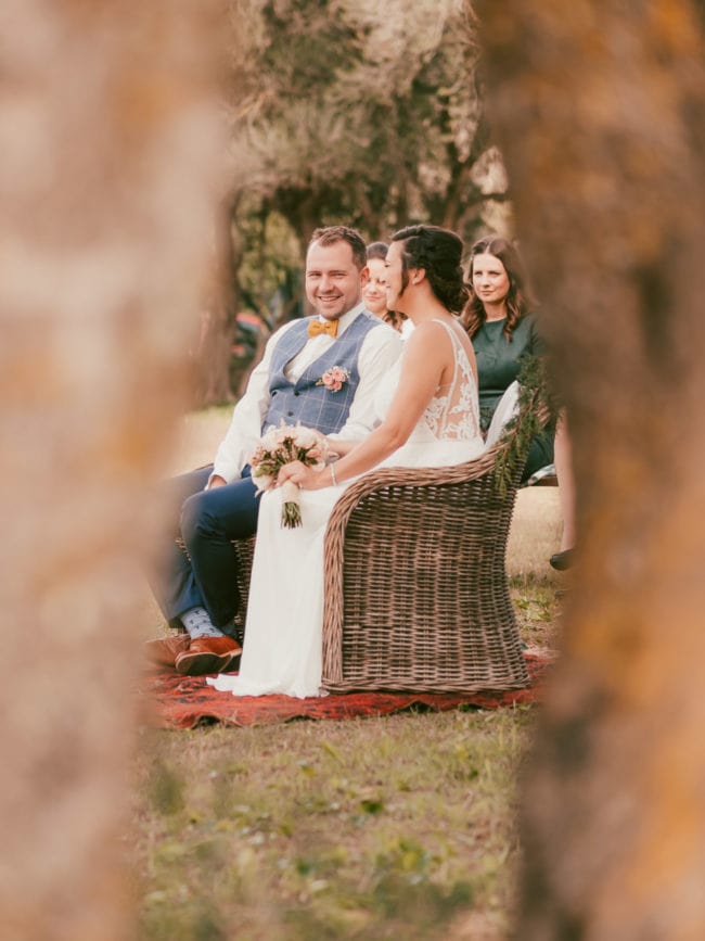 Eine Braut und ein Bräutigam sitzen lächelnd auf einer Korbbank im Freien, hinter ihnen sitzt eine Brautjungfer. Die Szene, die von einem Hochzeitsfotograf Mallorca aufgenommen wurde, wird von zwei unscharfen Baumstämmen im Vordergrund eingerahmt.
