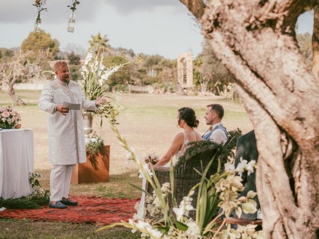 Ein Mann in traditioneller Kleidung steht im Freien und spricht mit einem sitzenden Paar während ihrer Hochzeit auf Mallorca. Das Paar sitzt auf einer Bank unter einem großen Baum, umgeben von Blumen und Grünzeug - wunderschön eingefangen von einem Hochzeitsfotograf Mallorca.