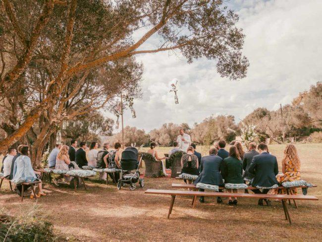 Eine Gruppe von Menschen sitzt auf Bänken im Freien unter Bäumen und nimmt an einer Hochzeit auf Mallorca teil. Der Trauzeuge steht vorne, während die Gäste nach vorne schauen, umgeben von Grün und einem teilweise bewölkten Himmel.