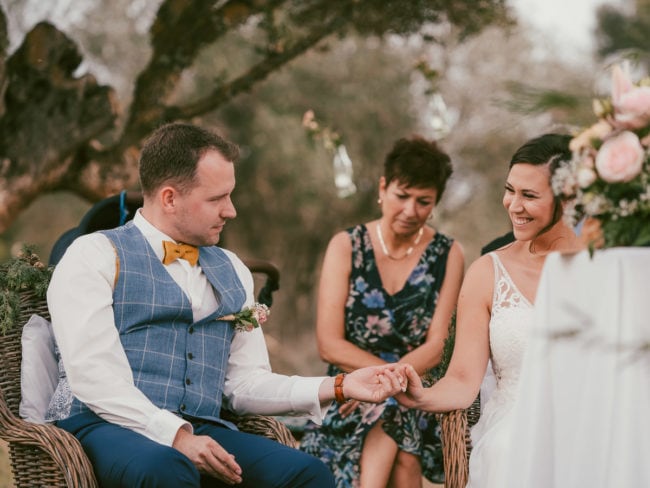 Braut und Bräutigam sitzen während ihrer Hochzeit auf Mallorca im Freien, lächelnd und händchenhaltend, mit einer Frau in einem Blumenkleid hinter ihnen sitzend. Bäume und Blumen tragen zu dem Charme bei, den ein Hochzeitsfotograf Mallorca eingefangen hat.