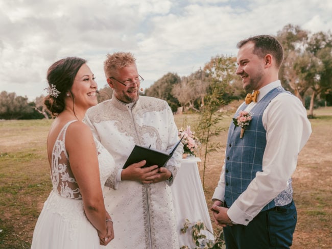 Eine Braut und ein Bräutigam lächeln sich während ihrer Hochzeit auf Mallorca an, während ein Geistlicher in einer weißen Robe aus einem Buch liest. Bäume und ein wolkenverhangener Himmel bilden die malerische Kulisse, wunderschön eingefangen von einem Hochzeitsfotograf Mallorca.